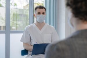 Profissional de saúde de máscara e uniforme branco, com prancheta, conversa com paciente em consultório